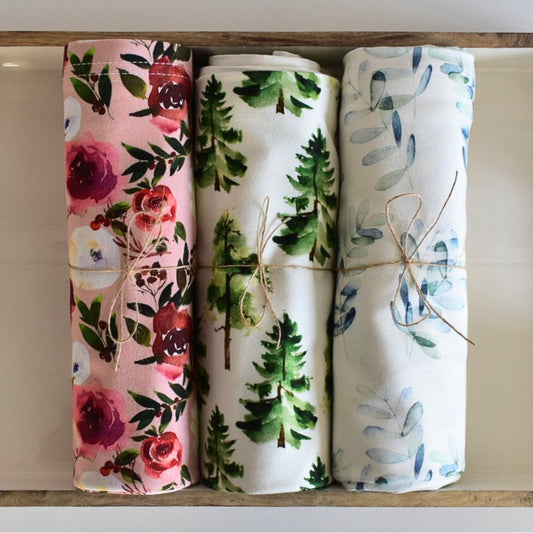 Soft rolled baby swaddles displayed in a wooden tray featuring three nature-inspired patterns: pink floral watercolor roses, watercolor evergreen trees, and soft blue eucalyptus leaves tied with rustic twine.